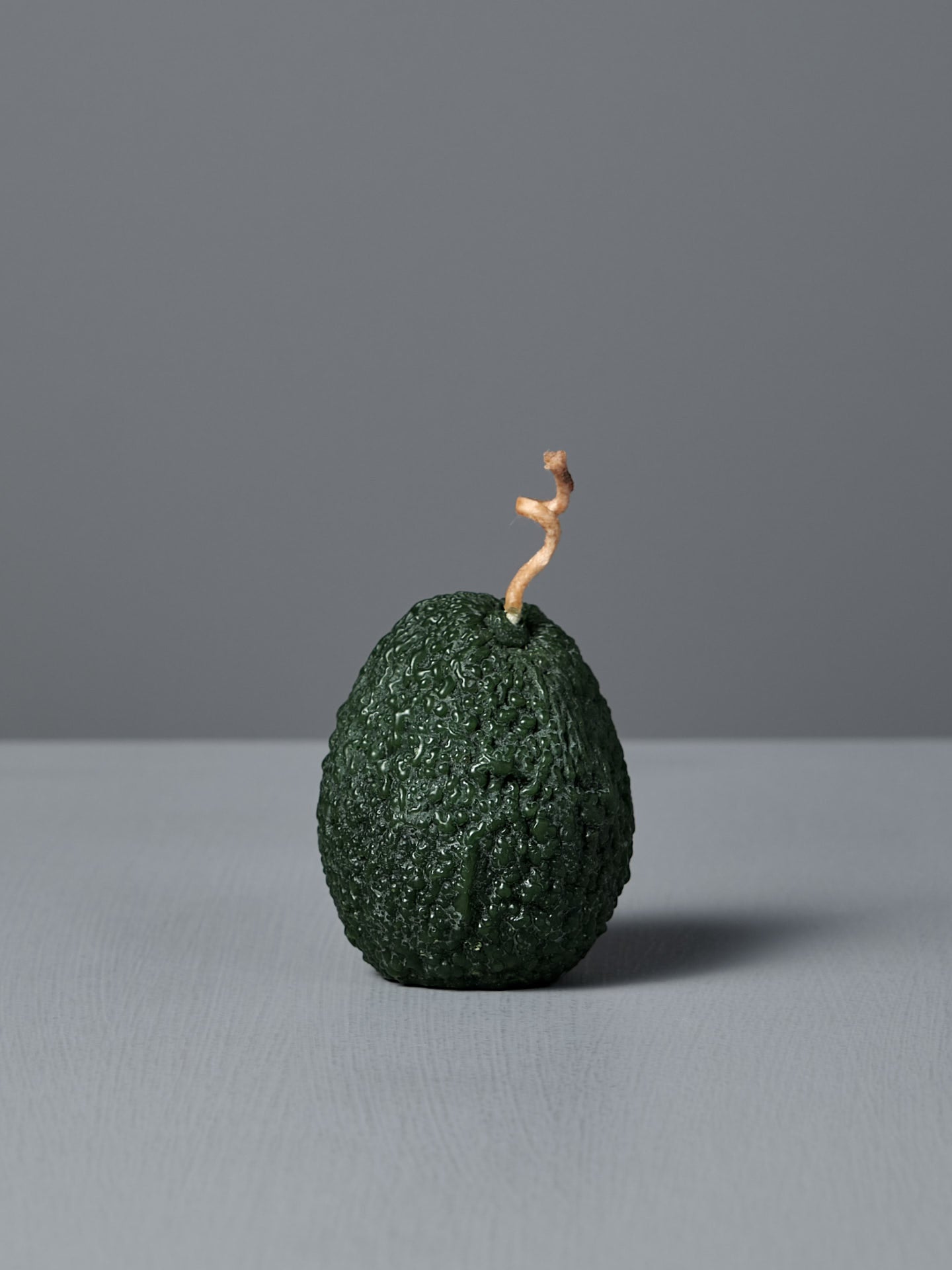 A light green, avocado-shaped sculptural candle called the Light Green Avocado Candle, made from pure beeswax with a short, twisted wick, sits on a light gray surface against a plain gray background.