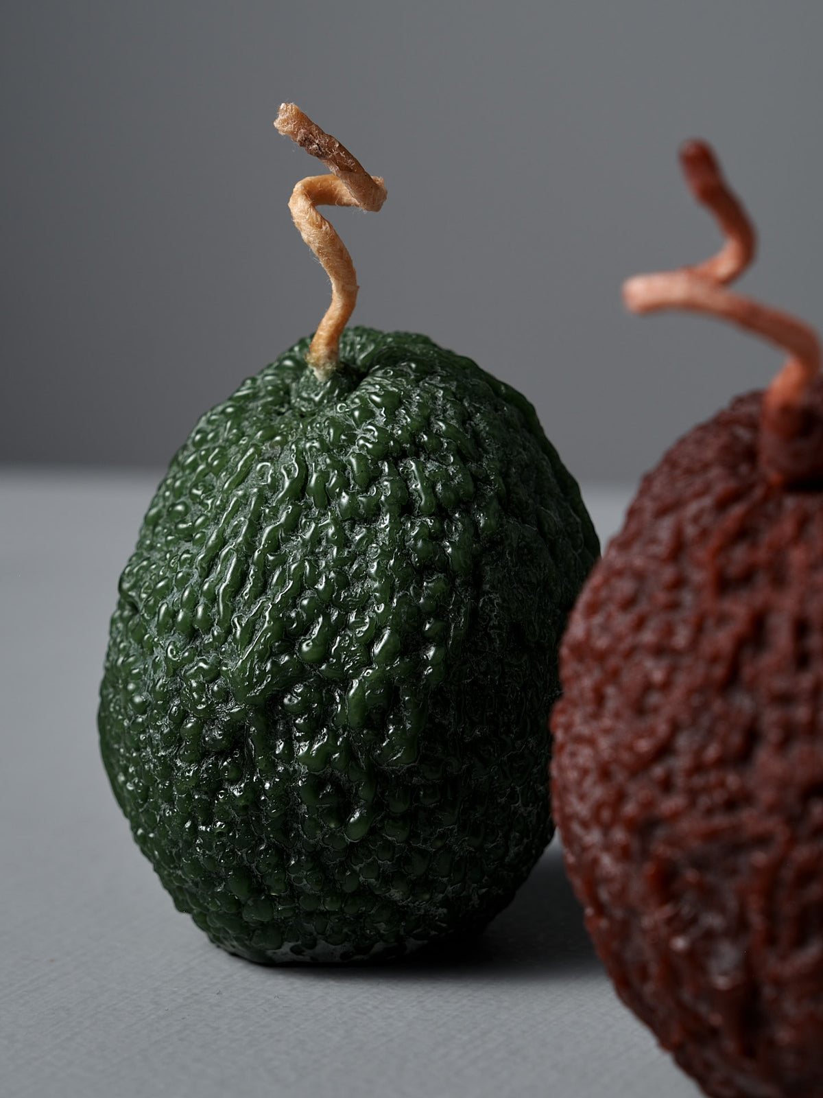 A Light Green Avocado Candle and a brown sculptural pure beeswax candle with twisted wicks, displayed against a neutral background.
