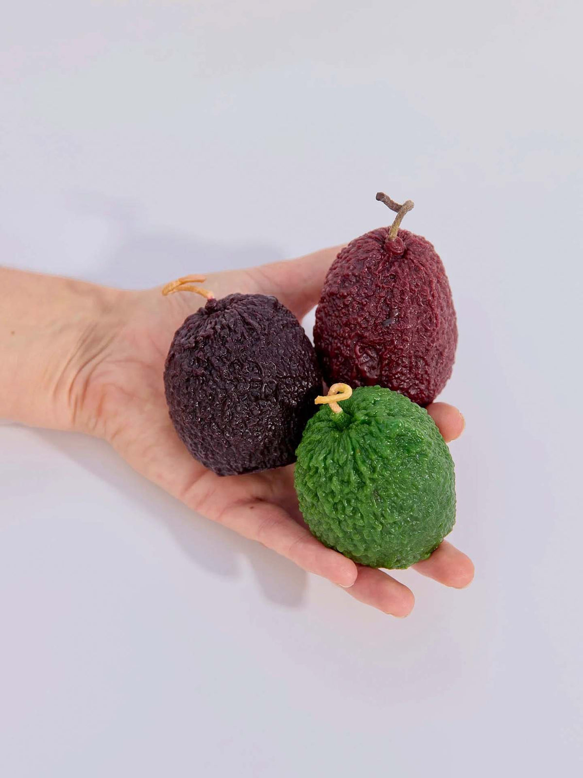 A hand holds three handmade beeswax candles shaped like avocados in green, deep purple, and reddish-purple hues—the Deep Purple Avocado Candle among them—crafted using New Zealand's candle-making traditions.