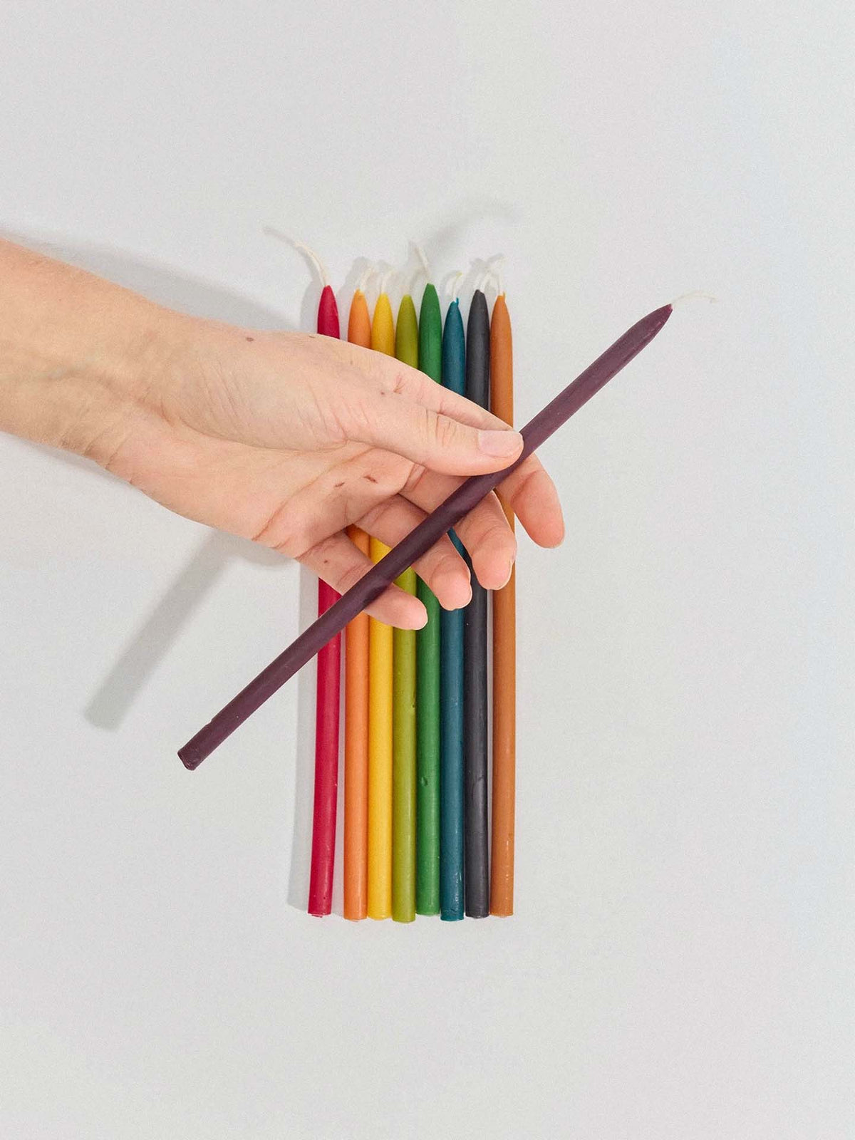 A hand holds a single dark purple Tall Celebration Candle – Mix & Match above a row of ten colorful taper candles neatly arranged on a white surface.