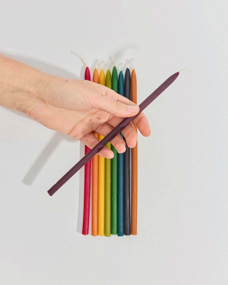 A hand holds a single dark purple Tall Celebration Candle – Mix & Match above a row of ten colorful taper candles neatly arranged on a white surface.