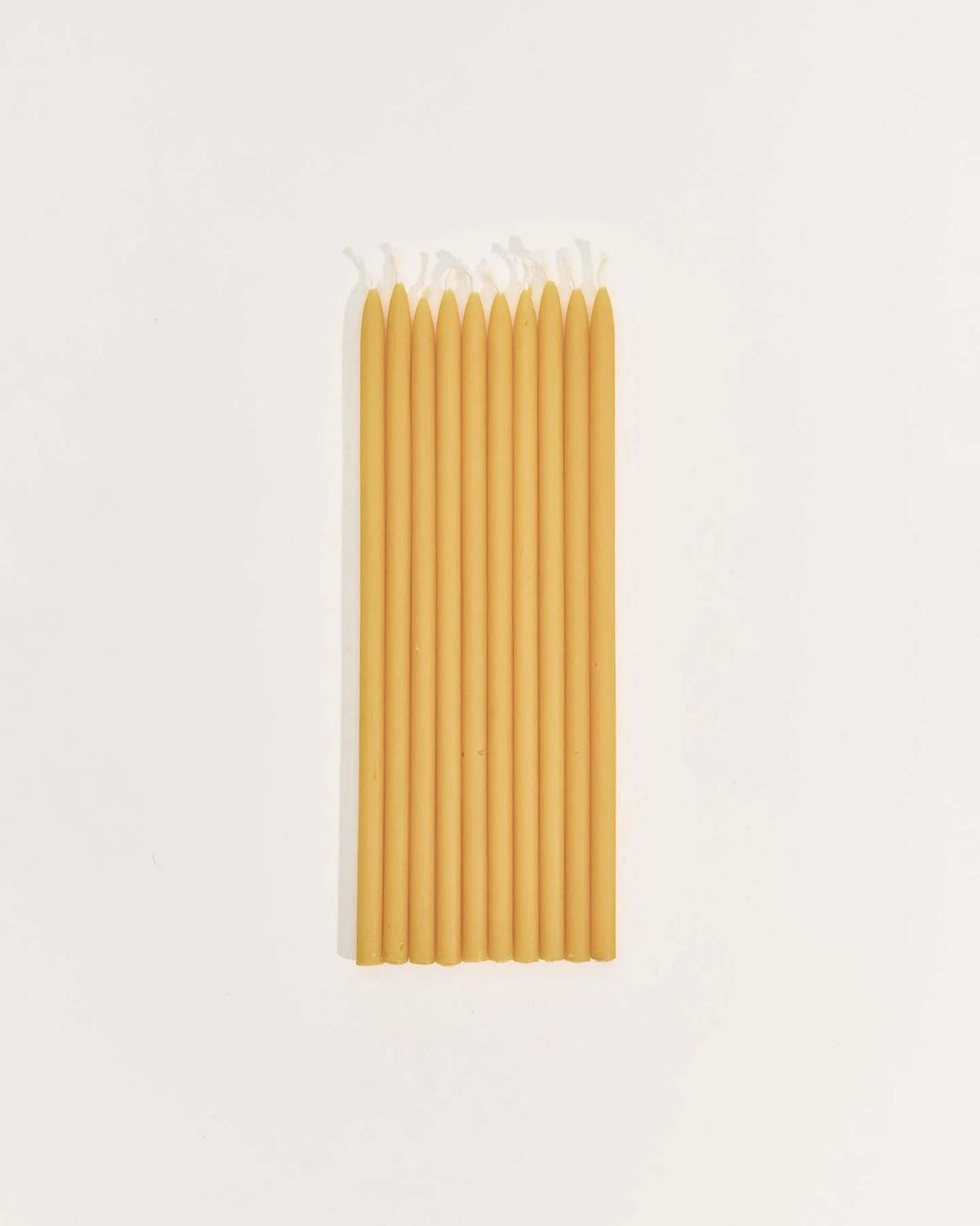 Nine tall, handmade beeswax Tall Celebration Candles – Mix & Match are arranged unlit in a neat row on a plain white surface.