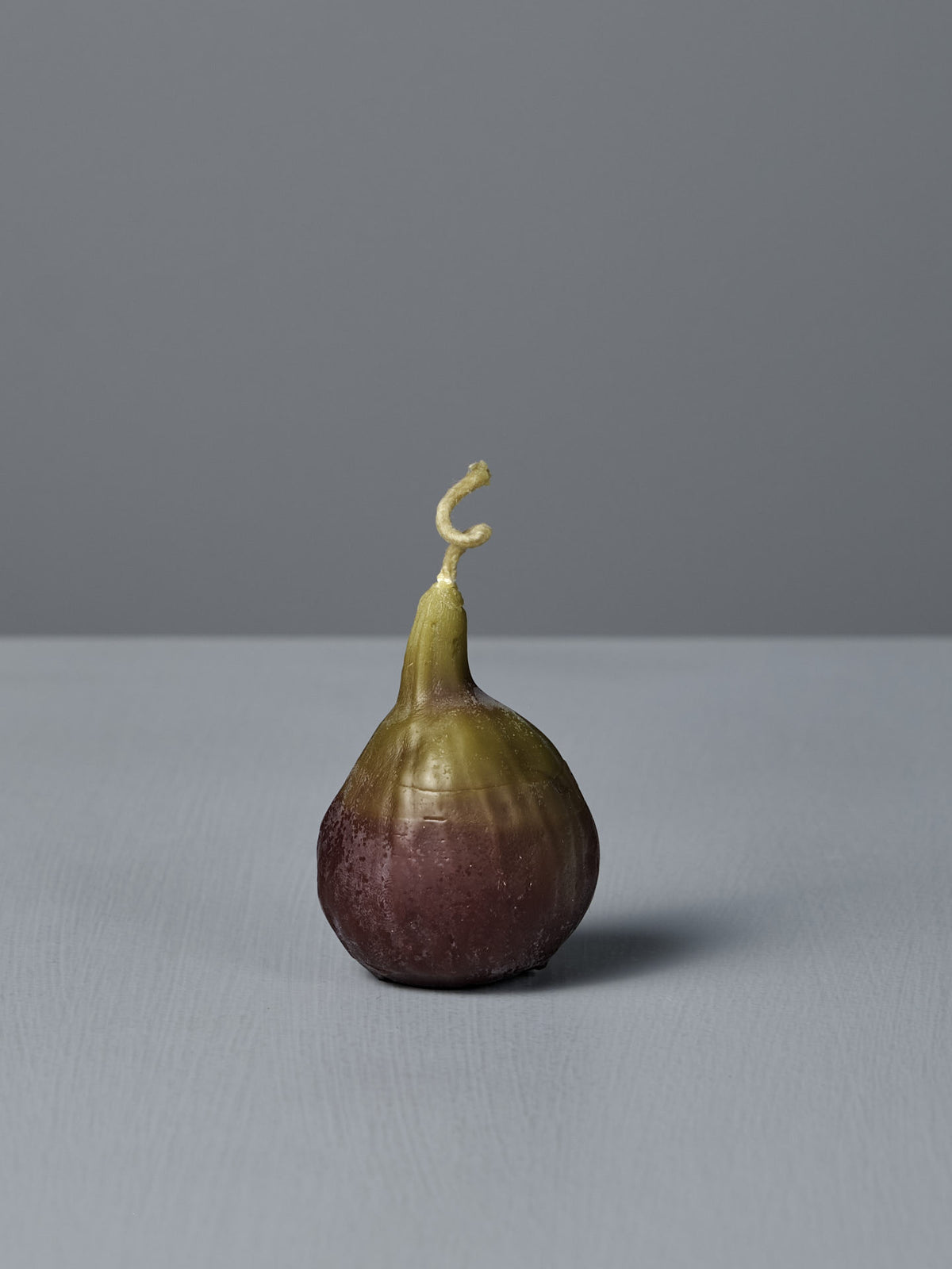 The Fig Candle, featuring a purple base and green top, stands upright on a light gray surface against a plain gray background, resembling a real fig.