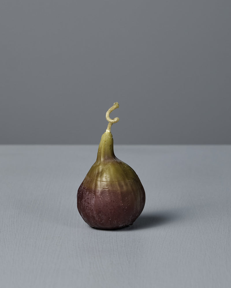 The Fig Candle, featuring a purple base and green top, stands upright on a light gray surface against a plain gray background, resembling a real fig.