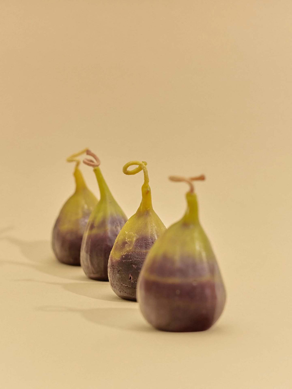 The Fig Candle is displayed beside four fresh figs with green and purple skin, all arranged on a beige surface against a plain beige background.