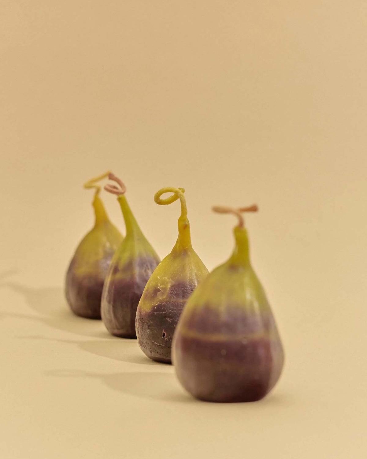 The Fig Candle is displayed beside four fresh figs with green and purple skin, all arranged on a beige surface against a plain beige background.