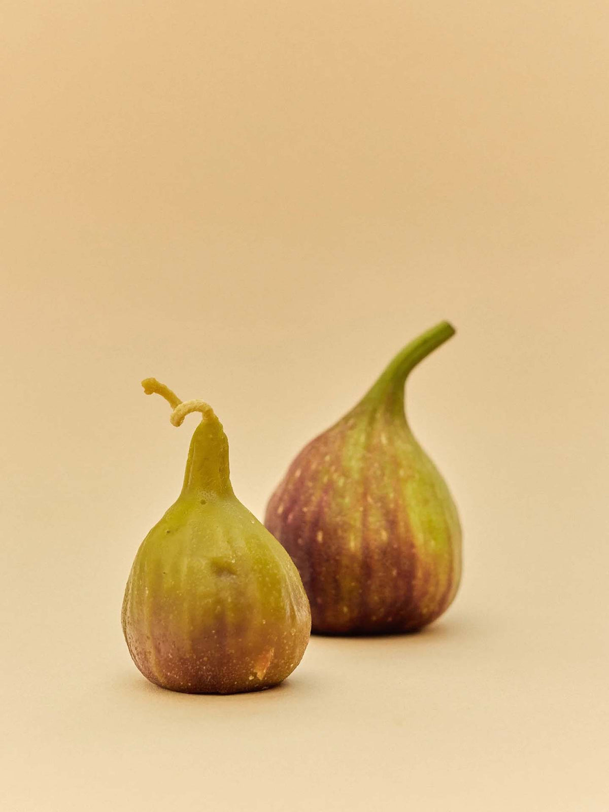 A handmade Fig Candle is displayed on a plain beige surface, accompanied by two fresh figs with green and purple hues, creating an inviting and natural scene.