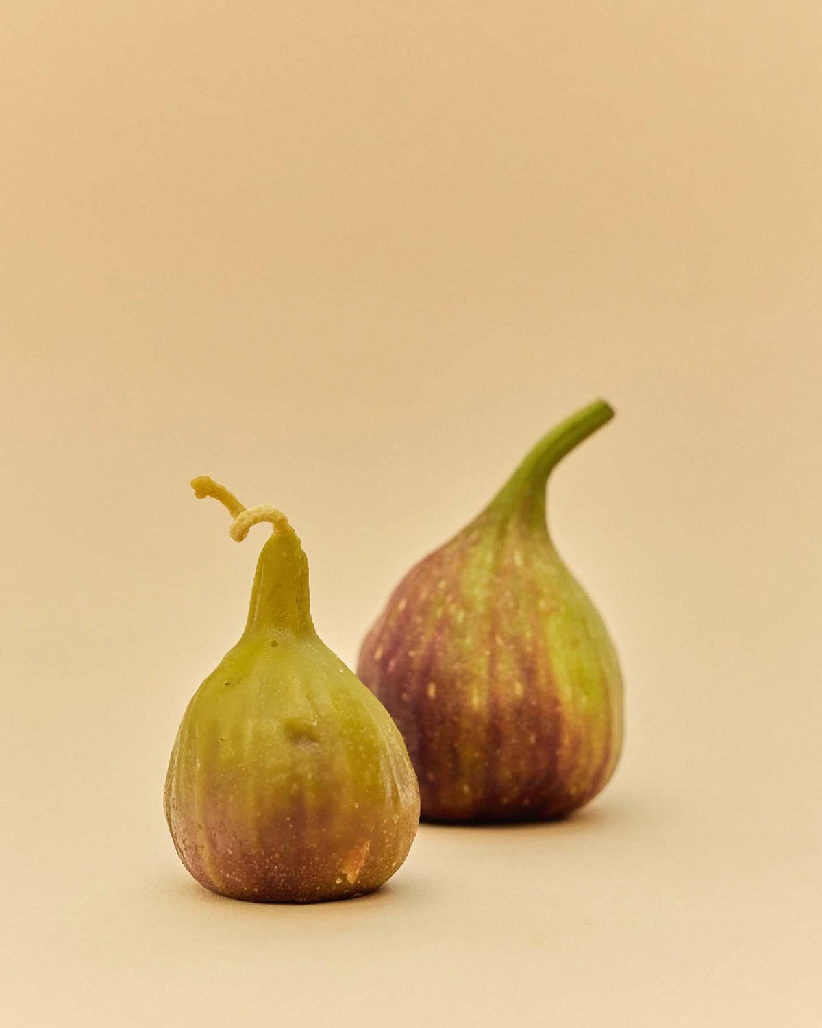 A handmade Fig Candle is displayed on a plain beige surface, accompanied by two fresh figs with green and purple hues, creating an inviting and natural scene.