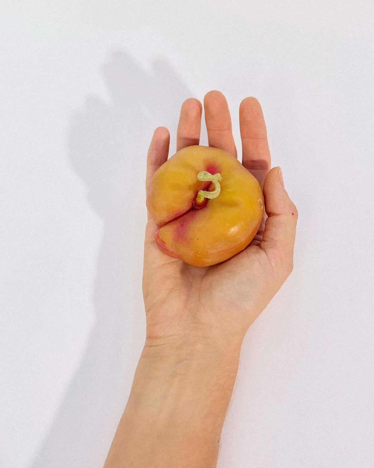 A hand holds a Flat Peach Candle with a misshapen yellow form and small green stem, resembling a real peach, against a plain white background.