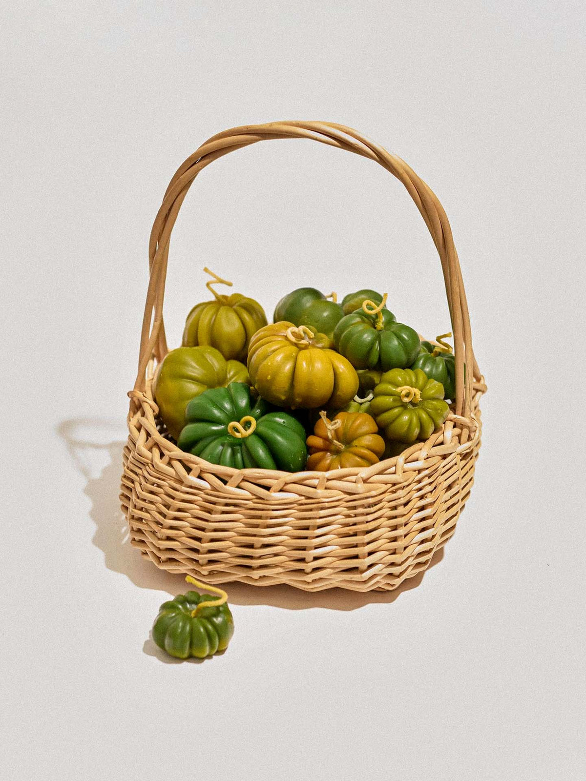 A wicker basket holds large Green Heirloom Tomato Candles, with one displayed outside the basket, all set against a plain white background.
