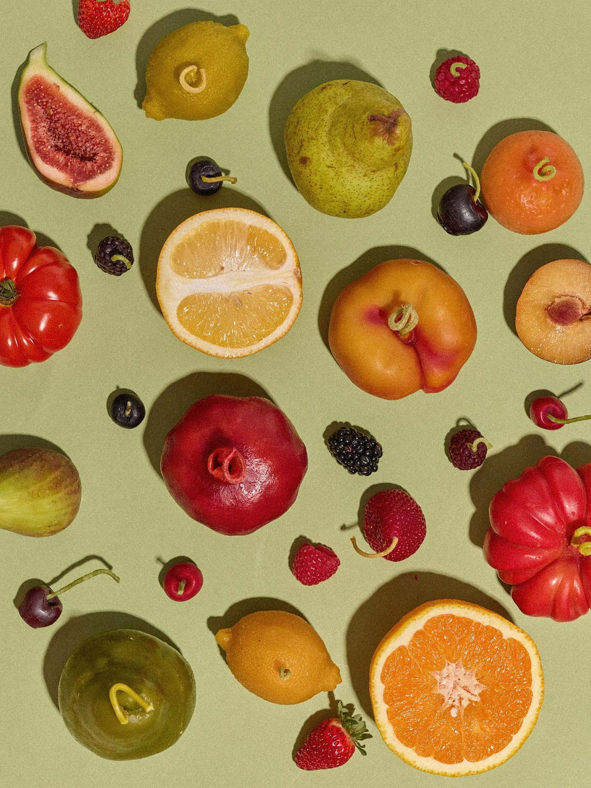 A selection of assorted fruits—figs, pears, citrus, cherries, and berries—arranged with an Orange Heirloom Tomato Candle (Small) on a light green background.