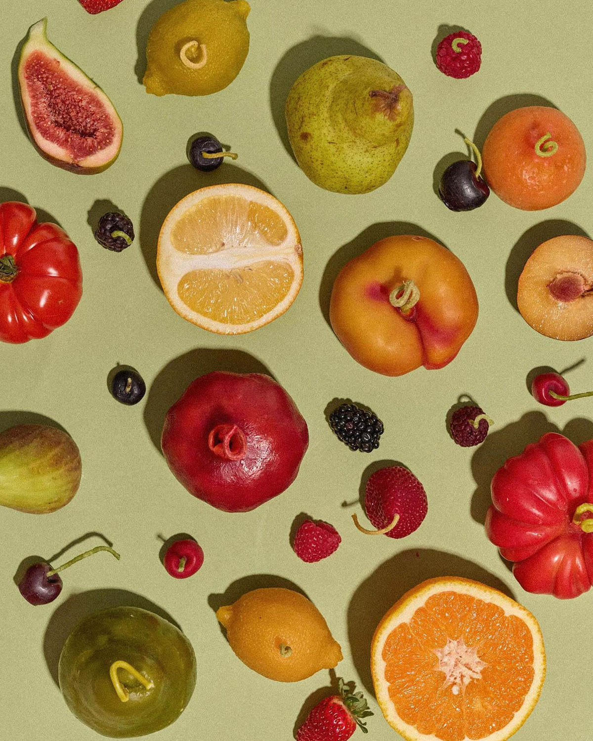 A selection of assorted fruits—figs, pears, citrus, cherries, and berries—arranged with an Orange Heirloom Tomato Candle (Small) on a light green background.
