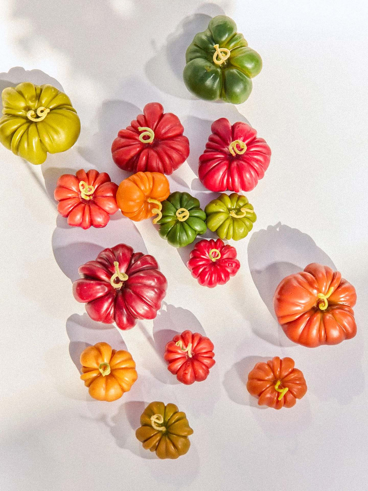 A selection of small pumpkins in orange, red, and green sits on a white surface with shadows, complemented by the Orange Heirloom Tomato Candle – Small to enhance the autumn atmosphere.