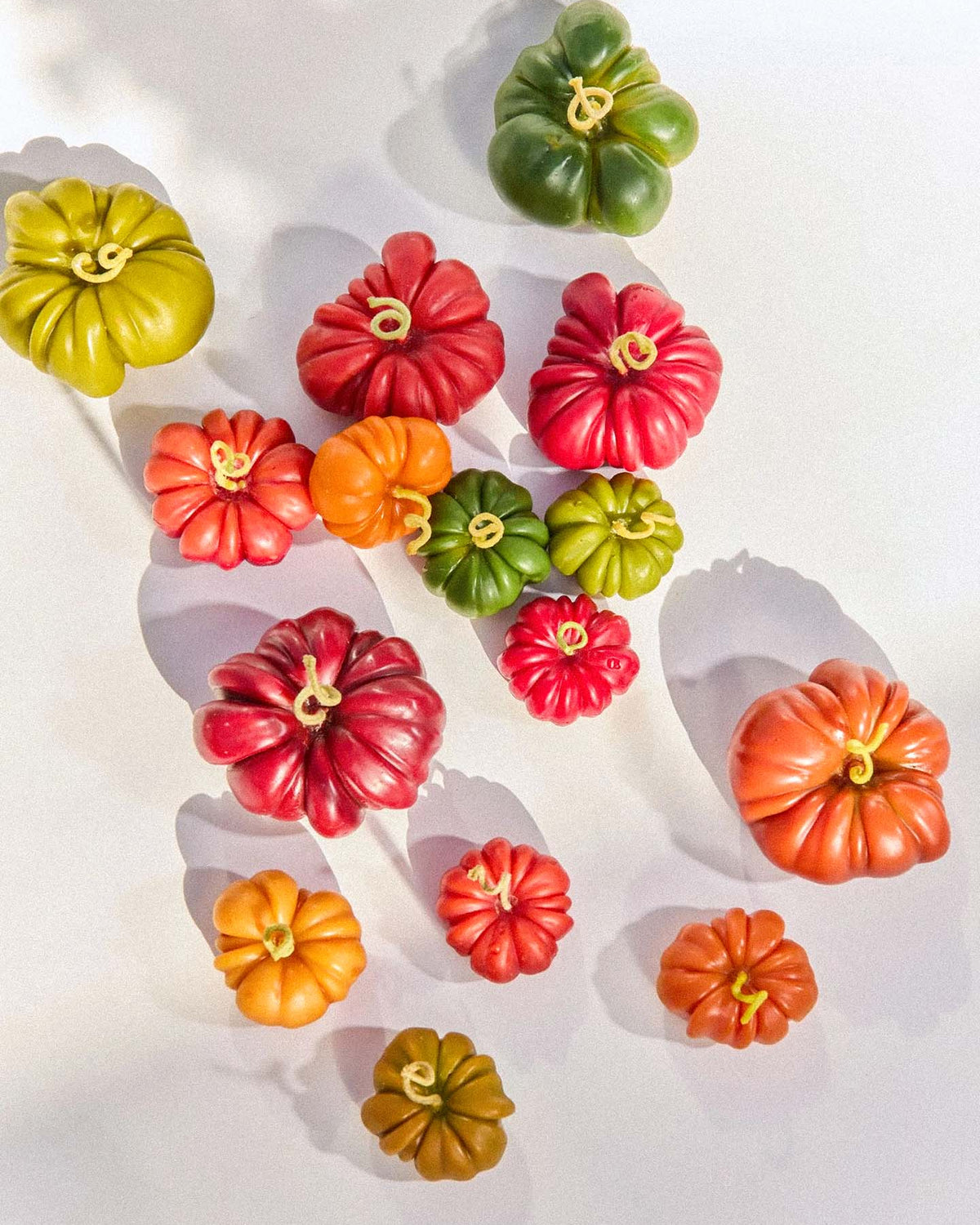 A selection of small pumpkins in orange, red, and green sits on a white surface with shadows, complemented by the Orange Heirloom Tomato Candle – Small to enhance the autumn atmosphere.