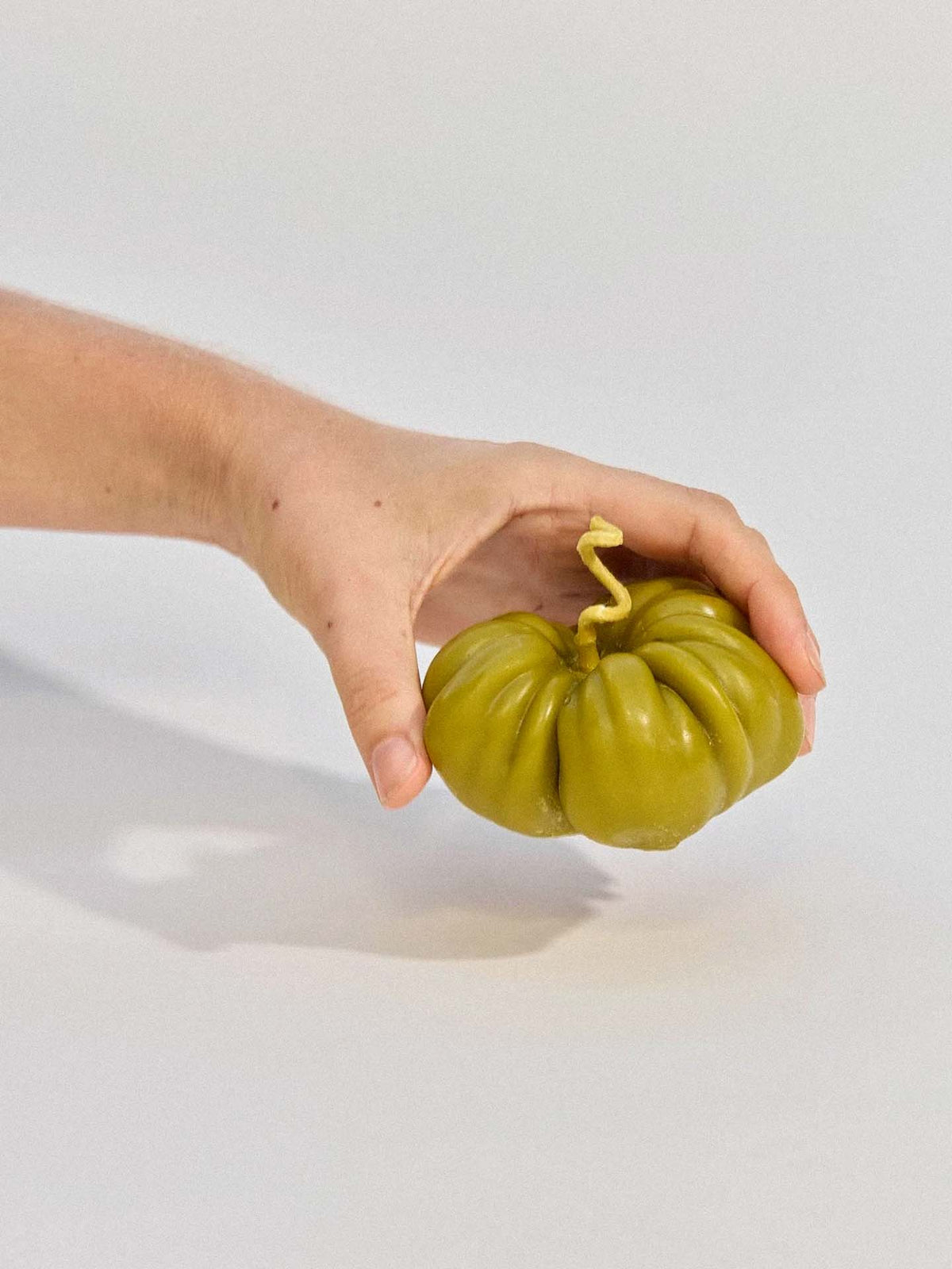 A hand holds the Green Heirloom Tomato Candle – Large, a green beeswax candle shaped like an heirloom tomato with a twisted stem, against a plain light background.
