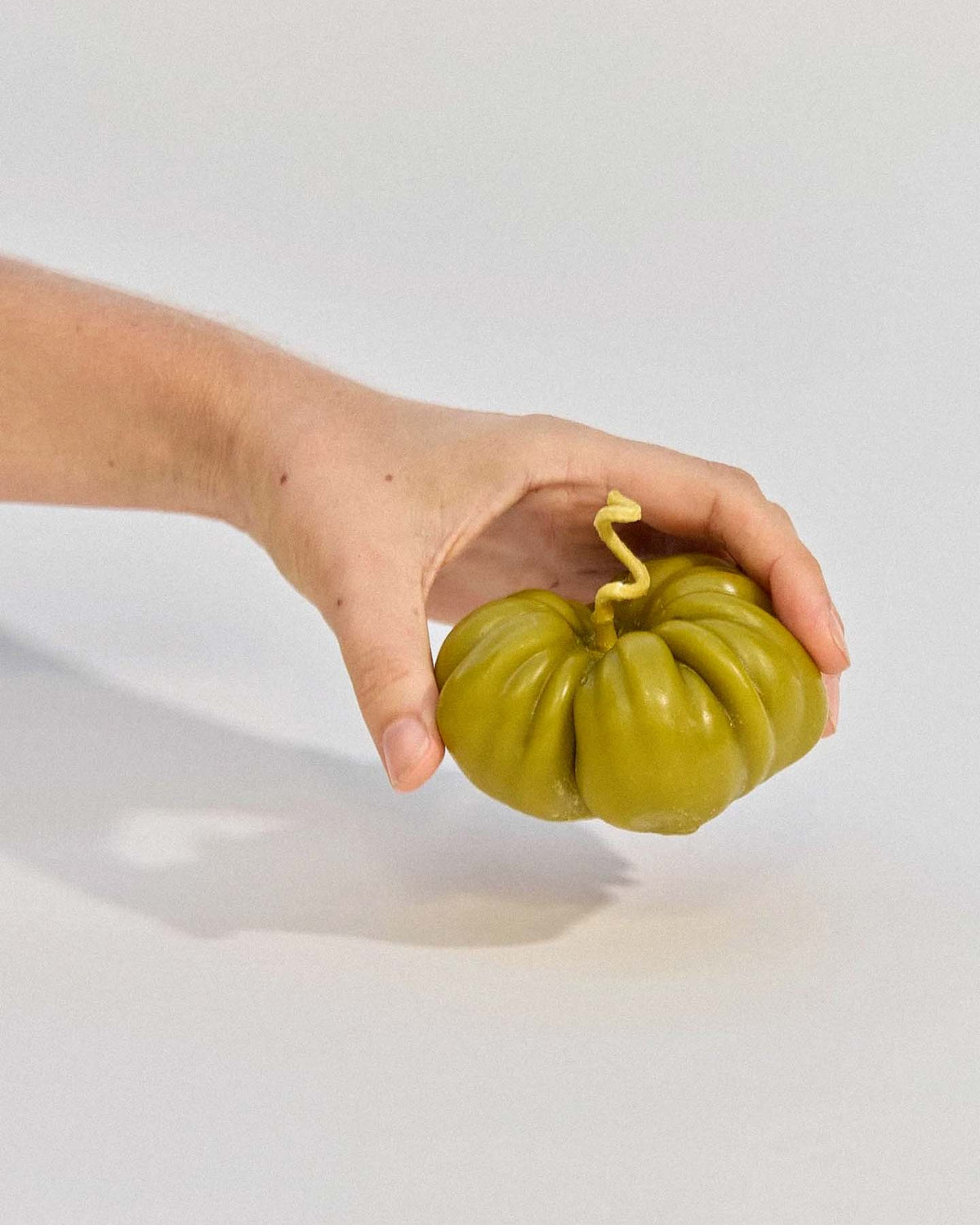 A hand holds the Green Heirloom Tomato Candle – Large, a green beeswax candle shaped like an heirloom tomato with a twisted stem, against a plain light background.