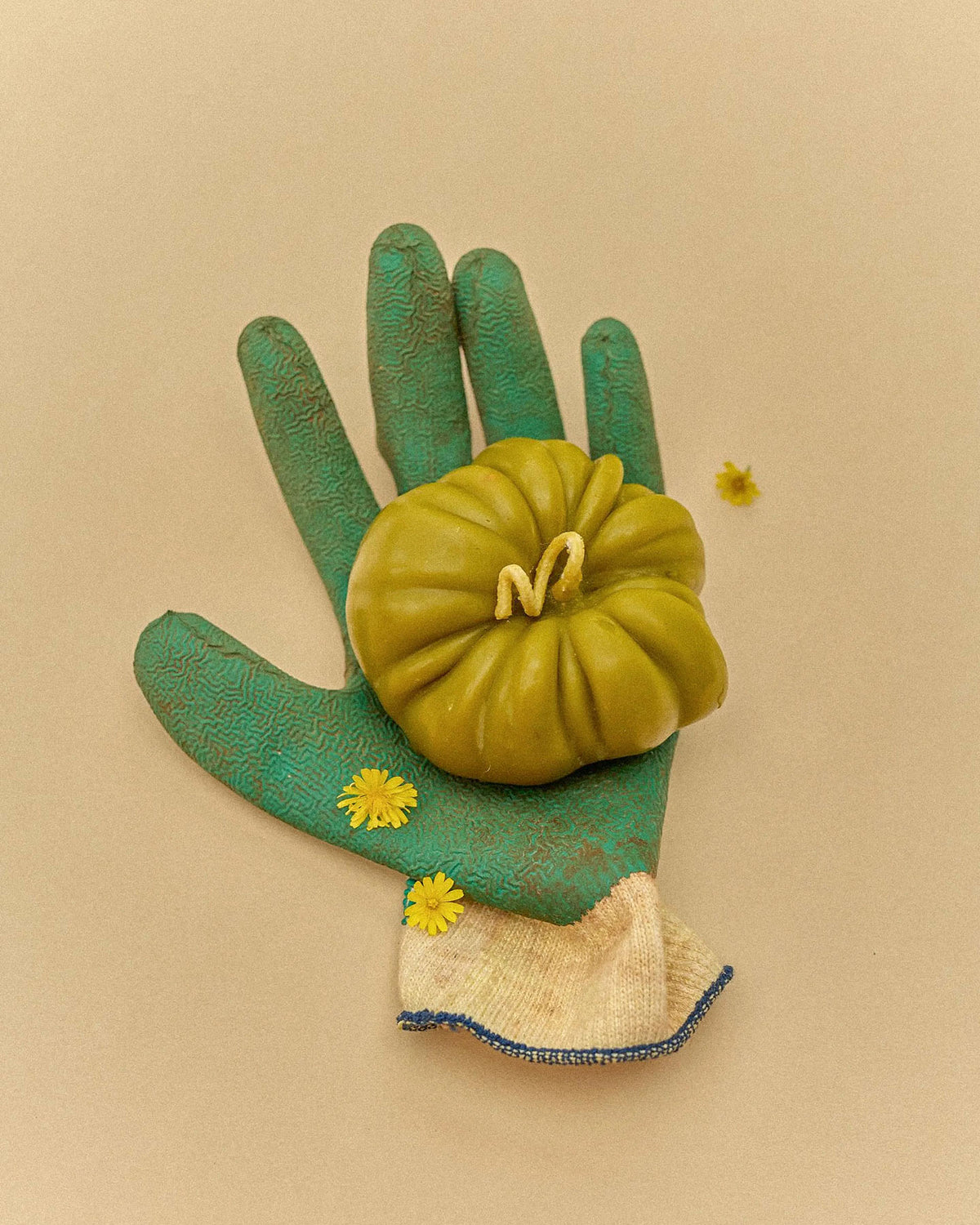 A green gardening glove holds a large Green Heirloom Tomato Candle, two yellow flowers, and a small green pumpkin, all arranged on its palm against a beige background.