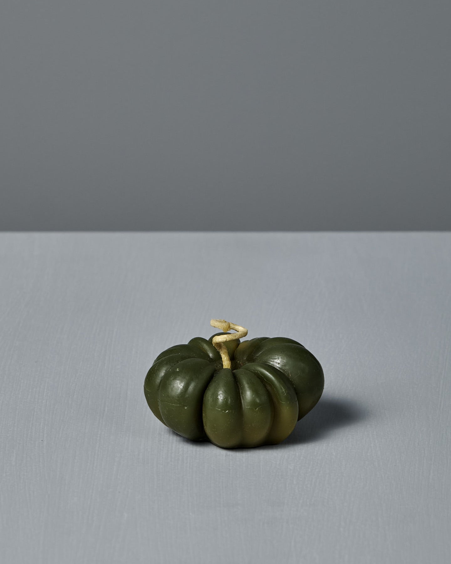 A small Green Heirloom Tomato Candle with a light stem sits on a flat, light gray surface, next to a handmade beeswax candle, all against a plain gray background.