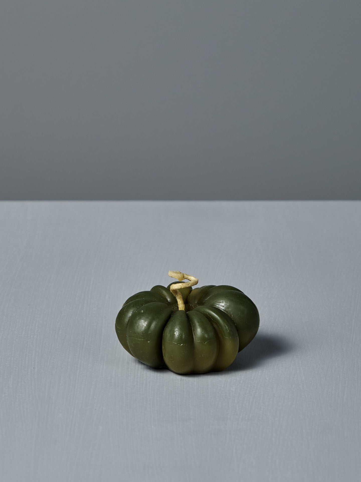 A small Green Heirloom Tomato Candle with a light stem sits on a flat, light gray surface, next to a handmade beeswax candle, all against a plain gray background.
