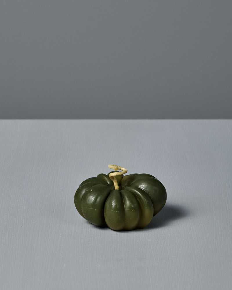 A small Green Heirloom Tomato Candle with a light stem sits on a flat, light gray surface, next to a handmade beeswax candle, all against a plain gray background.