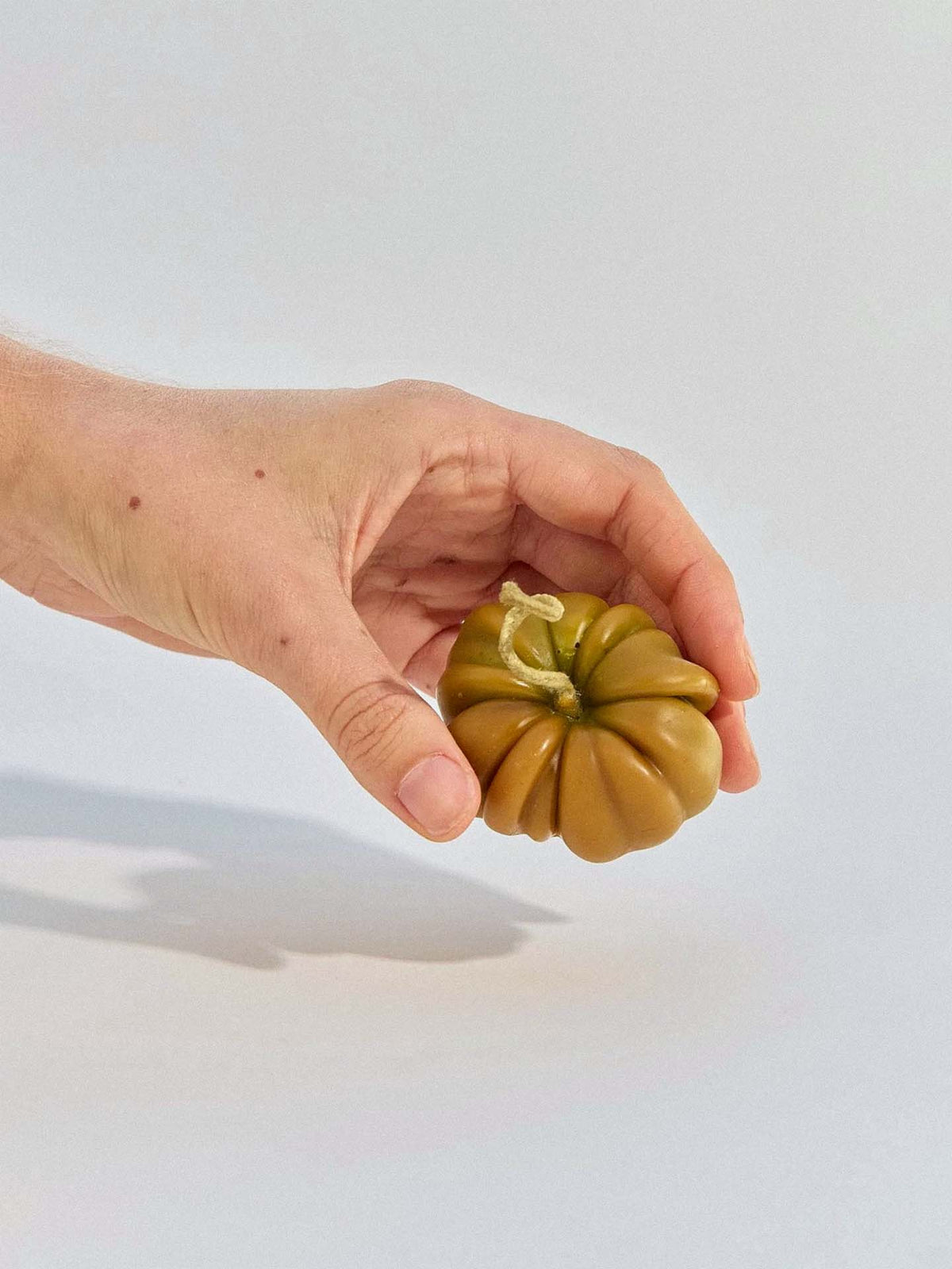 A hand holds the Green Heirloom Tomato Candle – Small, a greenish-brown, ribbed candle that resembles a tomato, against a plain light background.