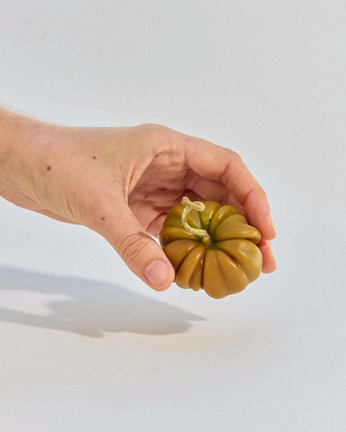A hand holds the Green Heirloom Tomato Candle – Small, a greenish-brown, ribbed candle that resembles a tomato, against a plain light background.