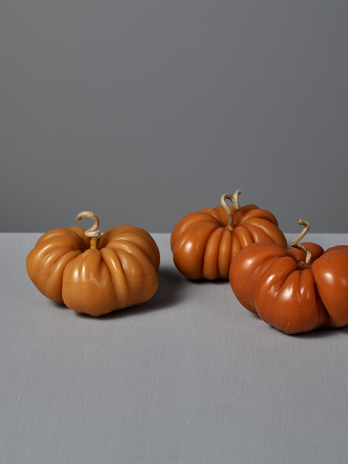 An Orange Heirloom Tomato Candle – Large is placed on a light grey surface alongside three decorative pumpkins with twisted stems, all set against a plain grey background.