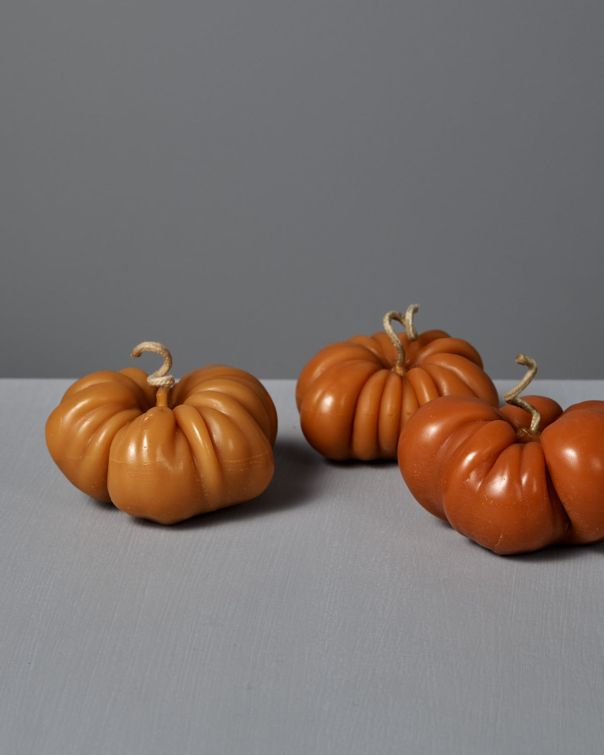 An Orange Heirloom Tomato Candle – Large is placed on a light grey surface alongside three decorative pumpkins with twisted stems, all set against a plain grey background.