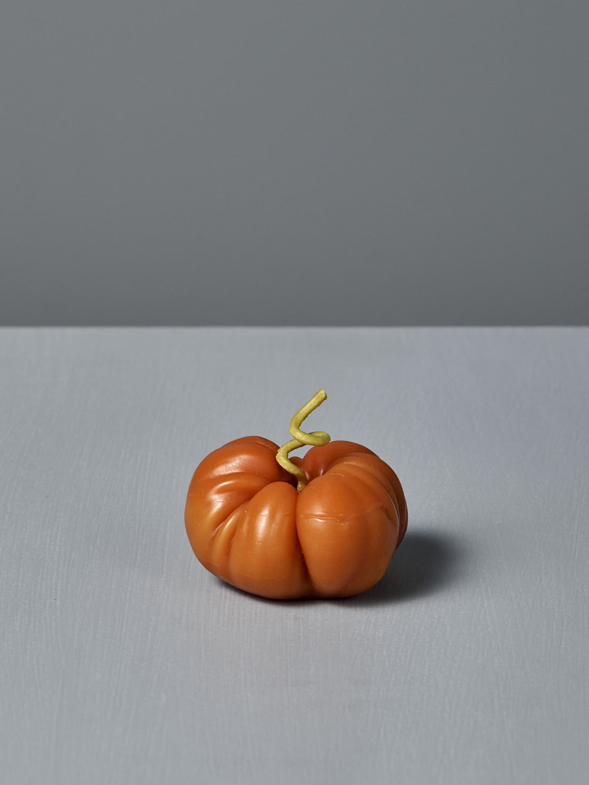 The Orange Heirloom Tomato Candle – Small features an irregular shape with a curly green stem, crafted by hand from premium New Zealand beeswax. It’s shown on a gray surface against a gray background.