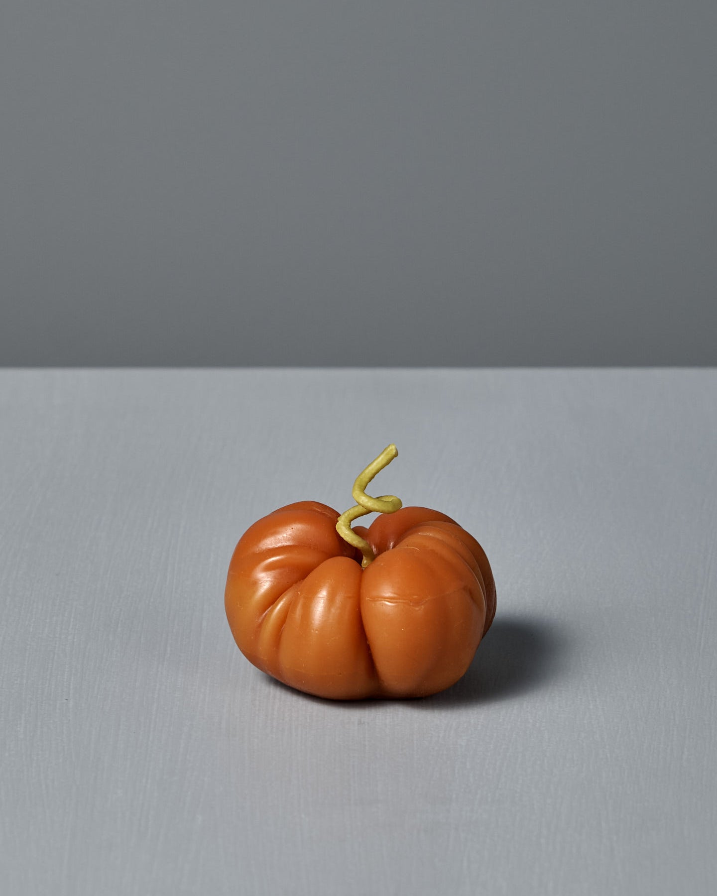 The Orange Heirloom Tomato Candle – Small features an irregular shape with a curly green stem, crafted by hand from premium New Zealand beeswax. It’s shown on a gray surface against a gray background.