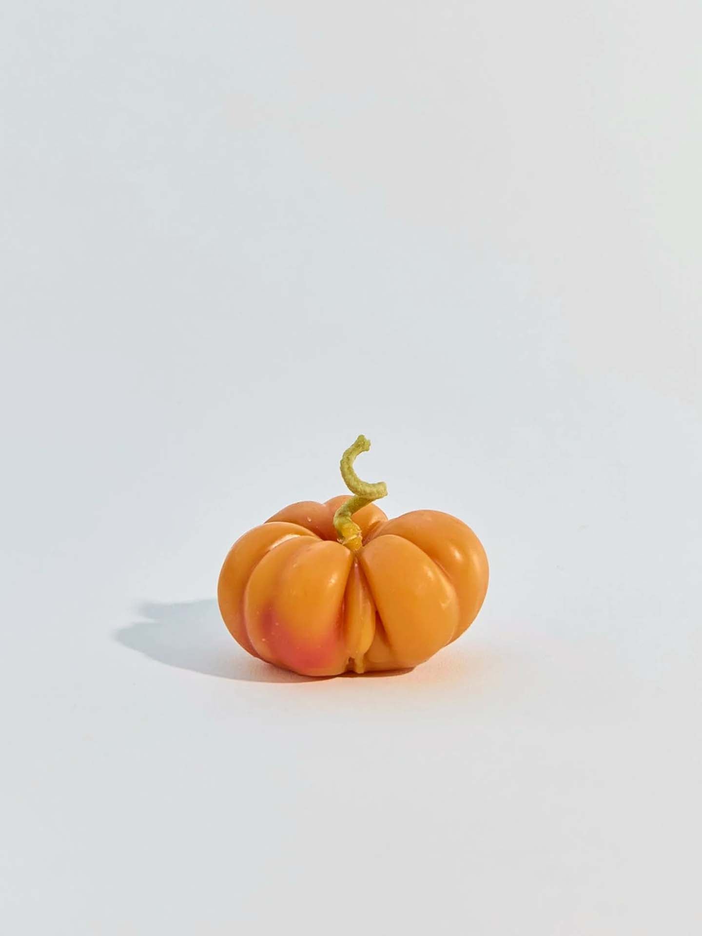 A small Orange Heirloom Tomato Candle with ridges and a short, curly stem rests on a plain white background beside a handmade New Zealand candle.