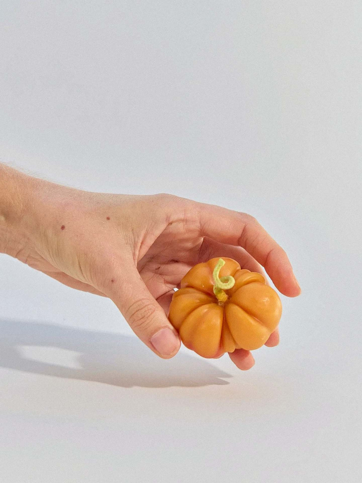 A hand holds the Orange Heirloom Tomato Candle – Small, a vibrant beeswax candle shaped like a ribbed tomato, against a plain light background.