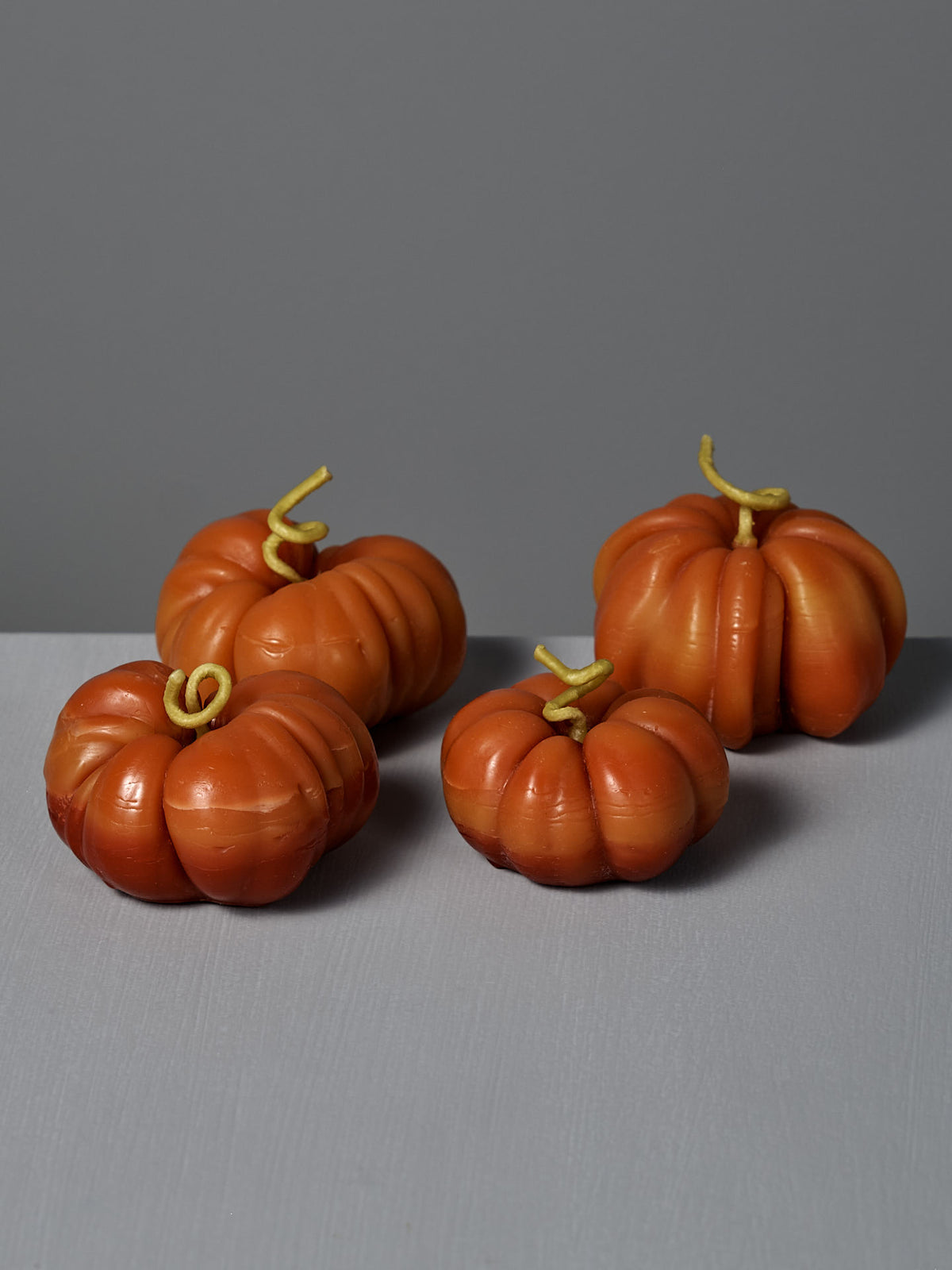 Four small Orange Heirloom Tomato Candles, each featuring a curled stem, are arranged on a gray surface against a gray background, evoking the cozy glow of handmade beeswax craftsmanship.