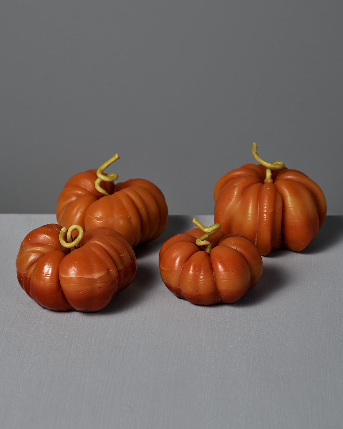 Four small Orange Heirloom Tomato Candles, each featuring a curled stem, are arranged on a gray surface against a gray background, evoking the cozy glow of handmade beeswax craftsmanship.