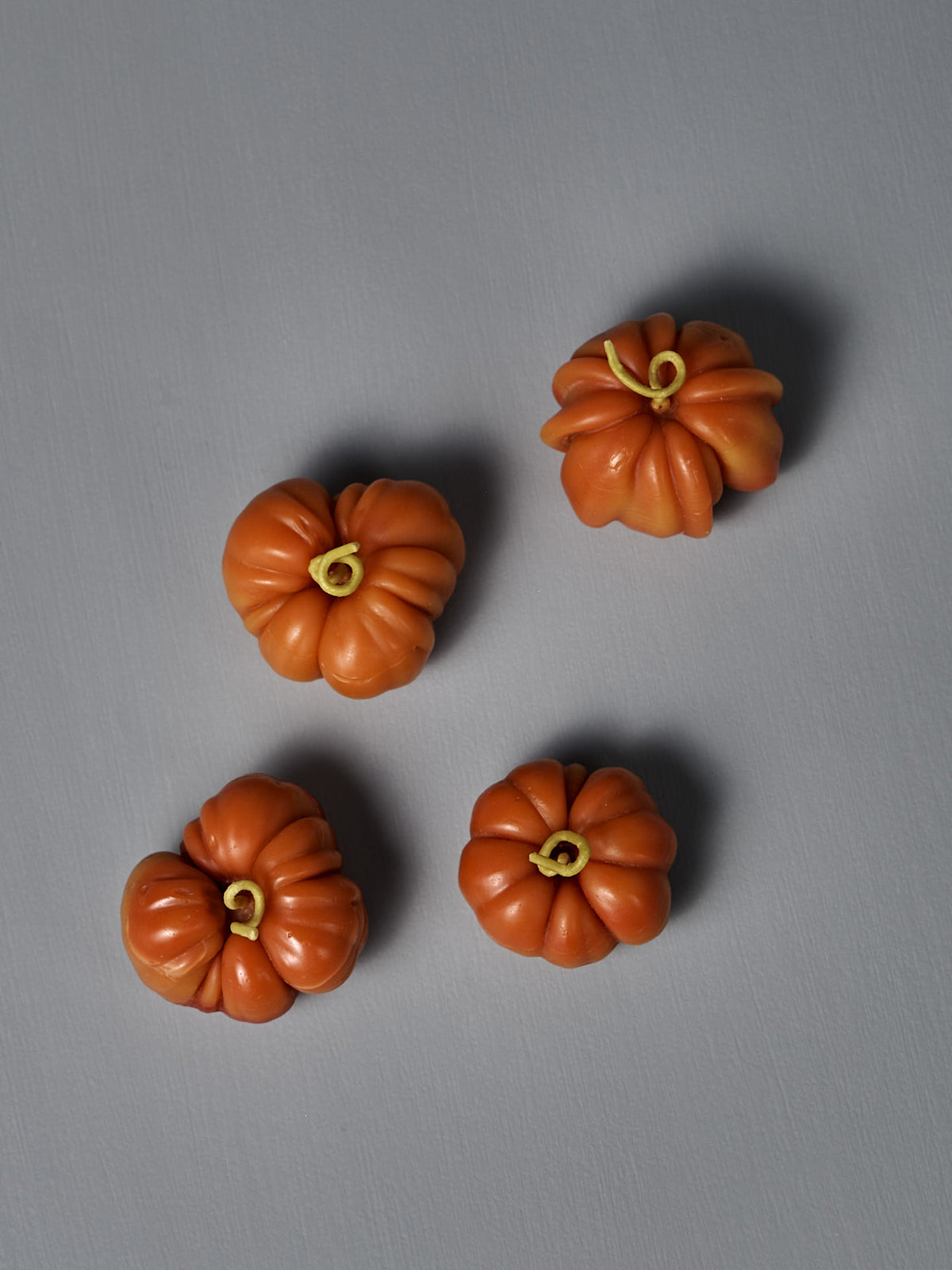 Four small, orange Orange Heirloom Tomato Candle – Small are arranged on a plain gray surface, each handmade from beeswax and featuring yellow stems.