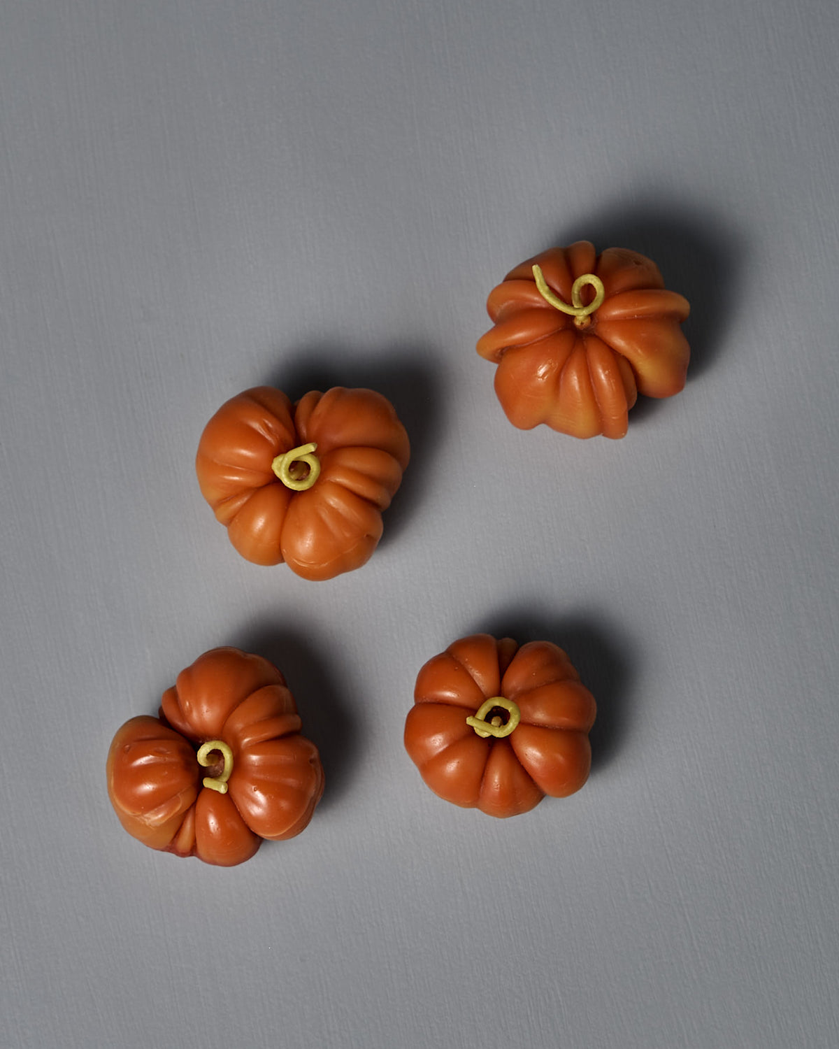 Four small, orange Orange Heirloom Tomato Candle – Small are arranged on a plain gray surface, each handmade from beeswax and featuring yellow stems.