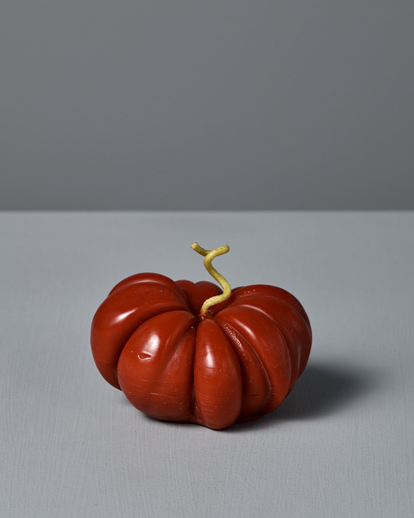 The large Red Heirloom Tomato Candle, featuring an irregular red shape and a short, curved yellow stem, sits on a plain light gray surface against a gray background—its unique appearance resembling a real heirloom tomato.