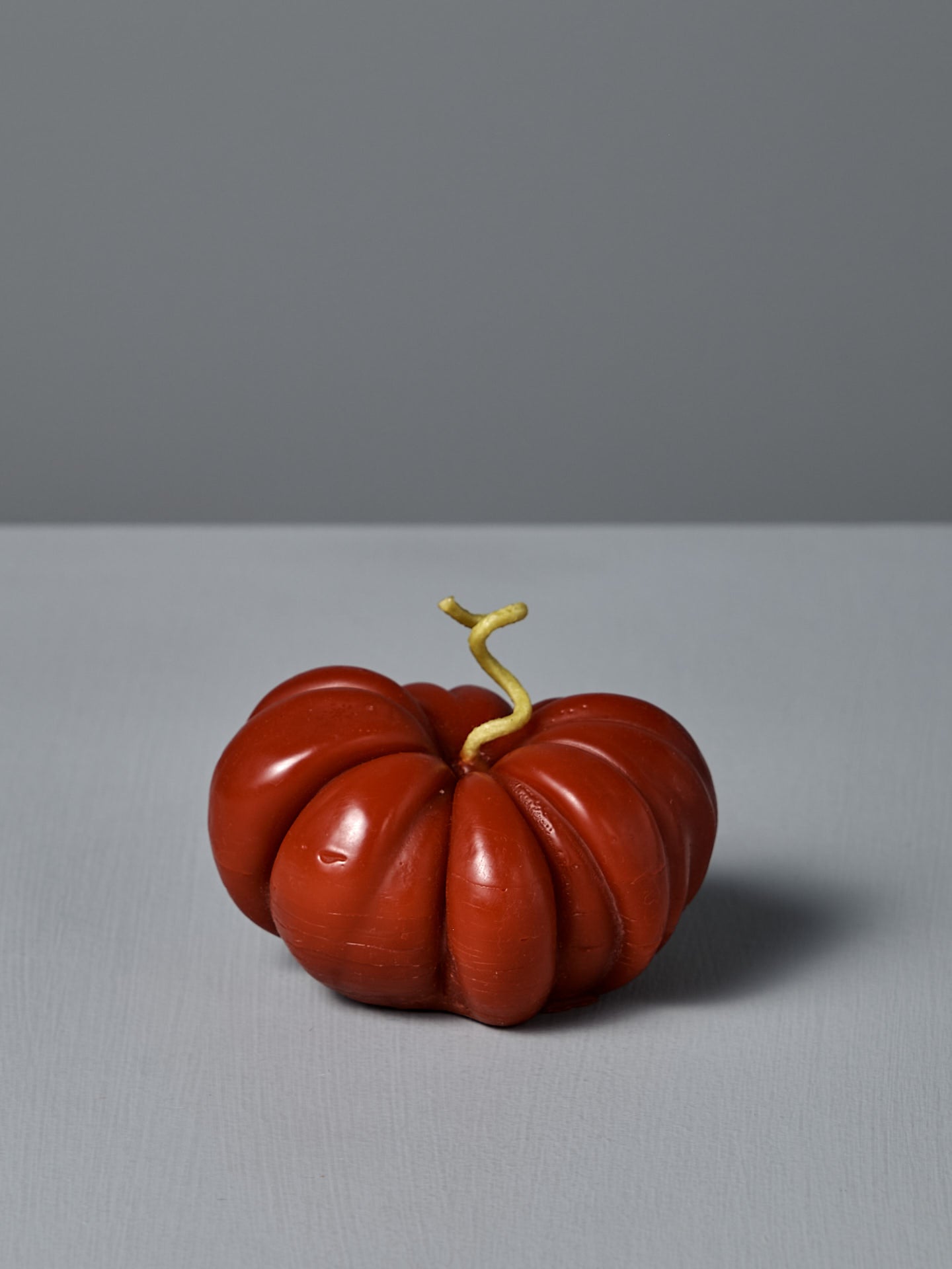 The large Red Heirloom Tomato Candle, featuring an irregular red shape and a short, curved yellow stem, sits on a plain light gray surface against a gray background—its unique appearance resembling a real heirloom tomato.