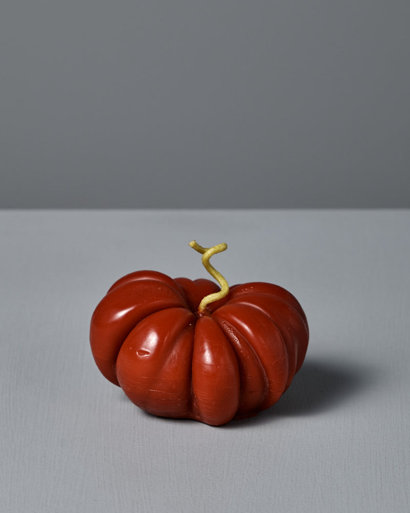 The large Red Heirloom Tomato Candle, featuring an irregular red shape and a short, curved yellow stem, sits on a plain light gray surface against a gray background—its unique appearance resembling a real heirloom tomato.