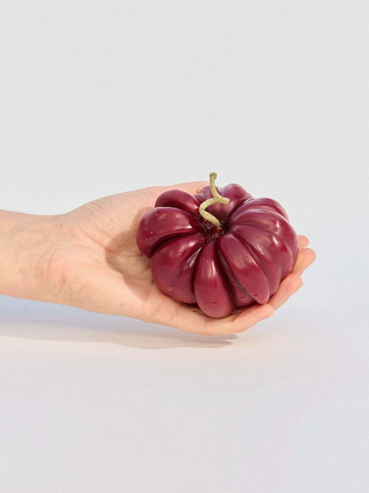 A hand holds the Red Heirloom Tomato Candle – Large, a uniquely ridged, handmade beeswax candle in deep red with a short green stem, crafted in New Zealand, set against a plain background.