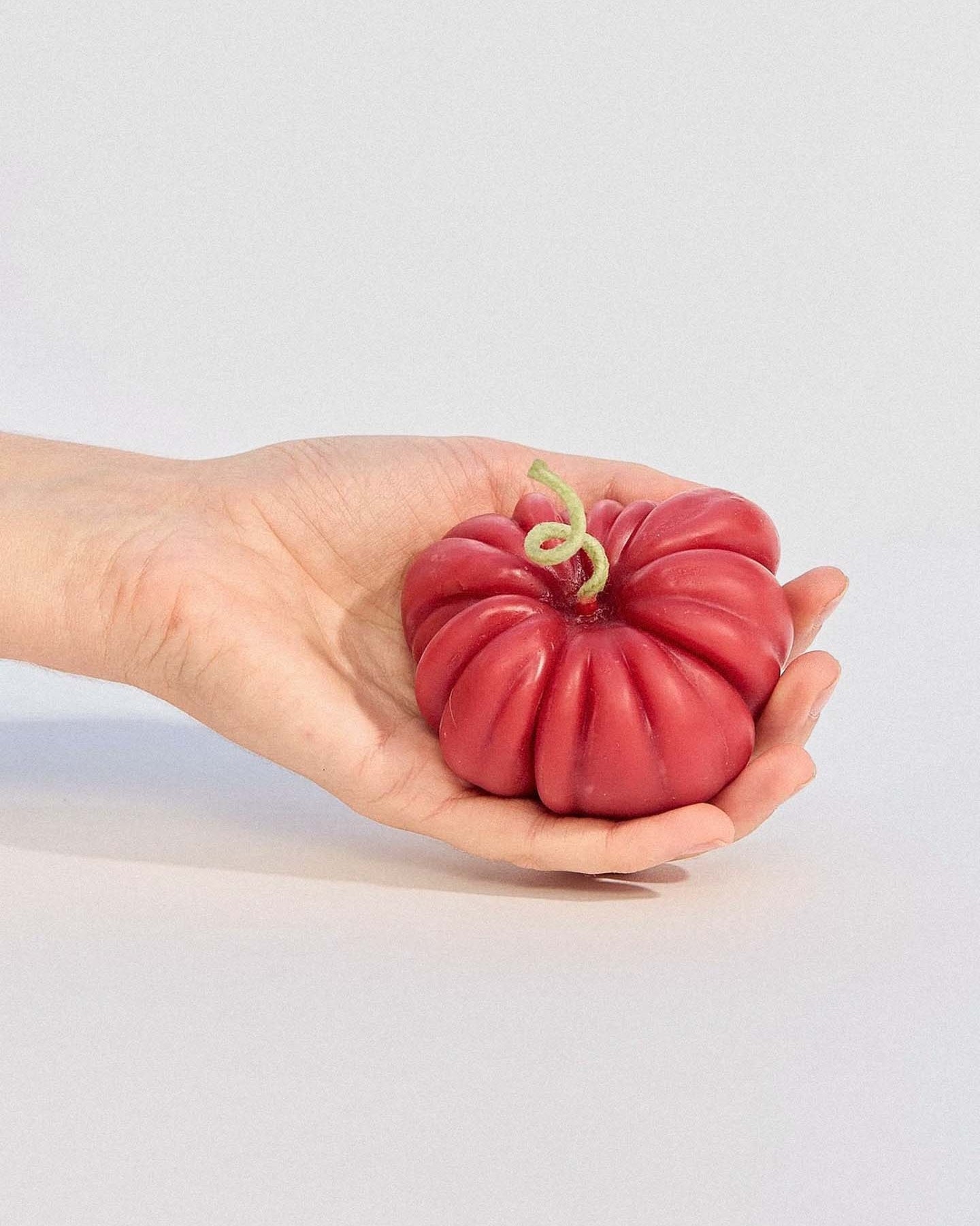 The large Red Heirloom Tomato Candle, featuring an irregular red shape and a short, curved yellow stem, sits on a plain light gray surface against a gray background—its unique appearance resembling a real heirloom tomato.
