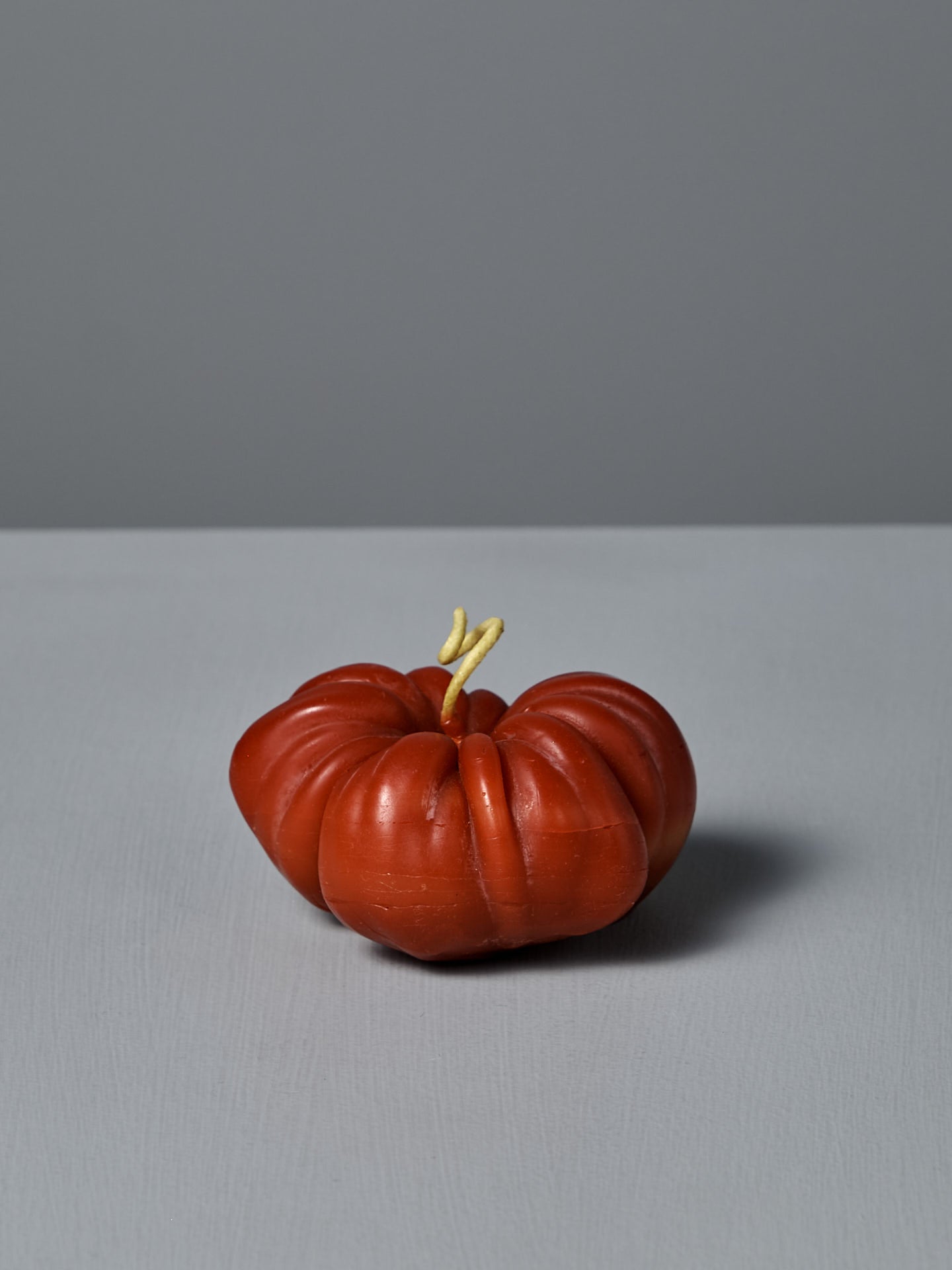 The Red Heirloom Tomato Candle – Small, designed with a realistic misshapen red form and a pale, forked stem, sits on a plain gray surface with a gray background.