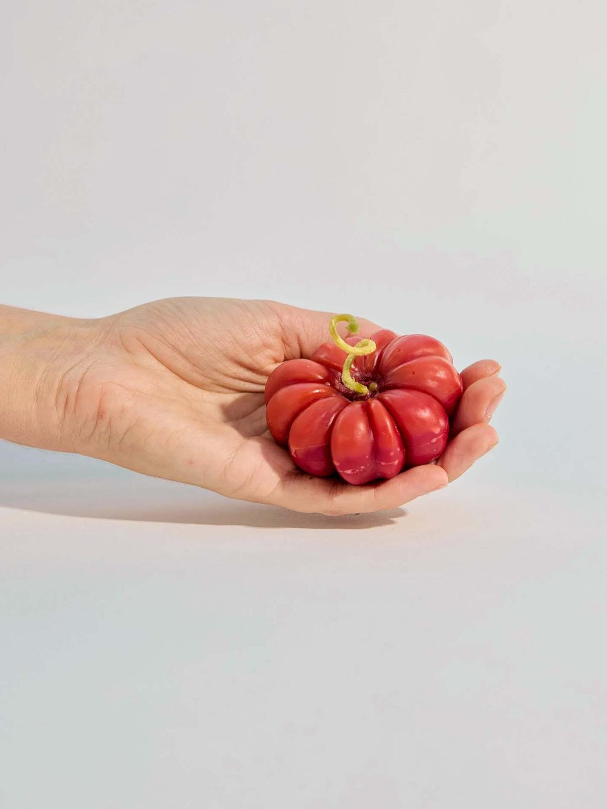 A hand holds the small Red Heirloom Tomato Candle, a flattened, ridged red candle with a green sprout, set against a plain light background.