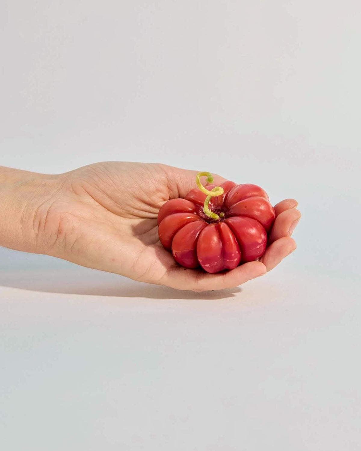 A hand holds the small Red Heirloom Tomato Candle, a flattened, ridged red candle with a green sprout, set against a plain light background.