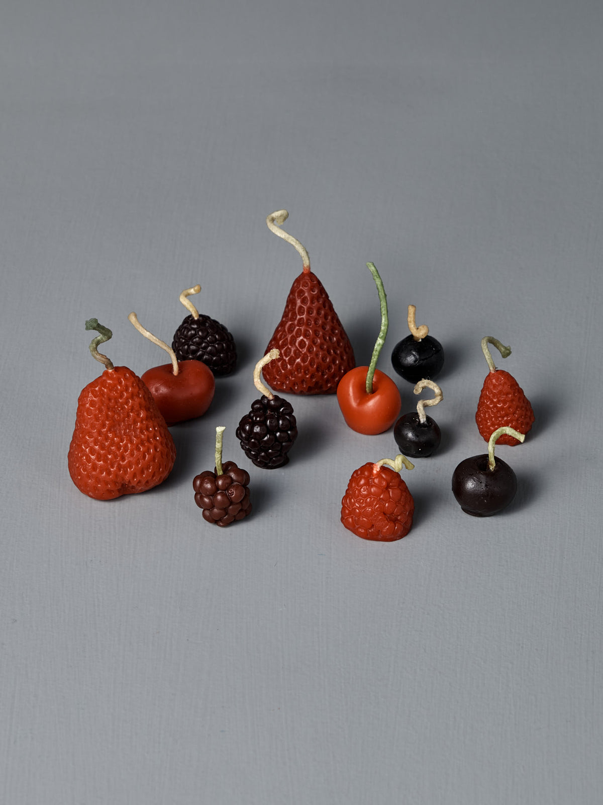 Mixed Berries Candles – Box of 10: Handmade beeswax candles shaped like strawberries, blackberries, cherries, raspberries, and grapes with twisted stems are arranged on a plain gray surface.