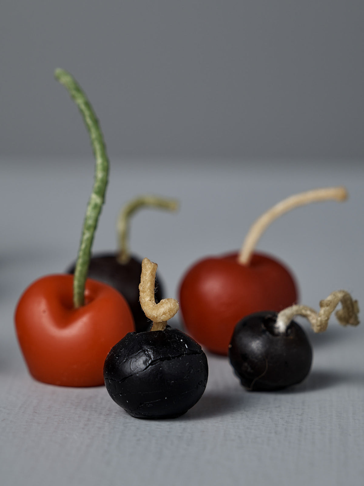 The Mixed Berries Candles – Box of 10 features four artificial cherries—two red and two dark purple—with assorted stems, resembling beeswax fruit candles, all displayed on a gray surface with a neutral backdrop.