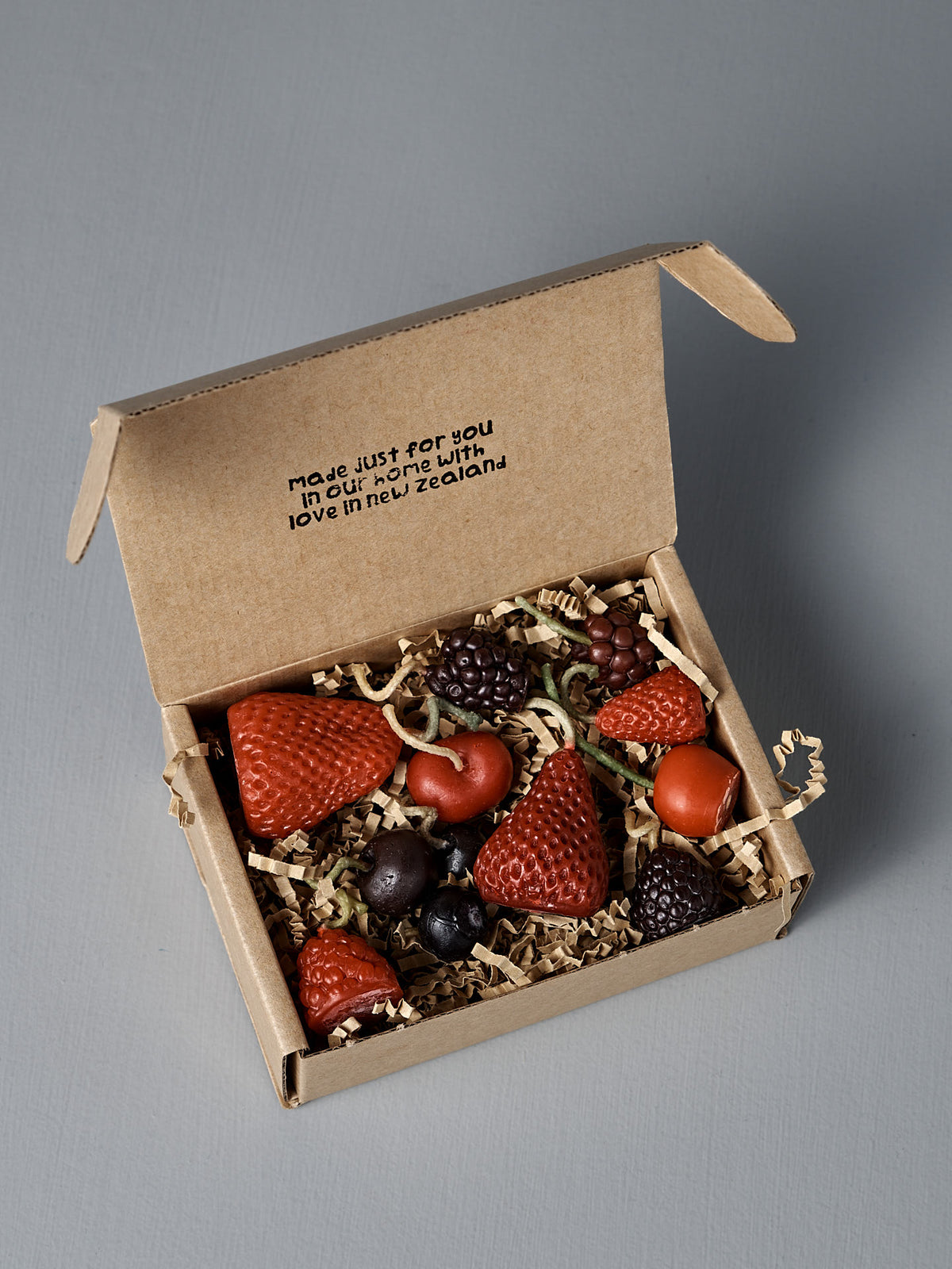 A small open cardboard box contains ten handmade Mixed Berries Candles shaped like fruit, nestled on shredded paper. The lid says, "Made just for you in our home with love in New Zealand.