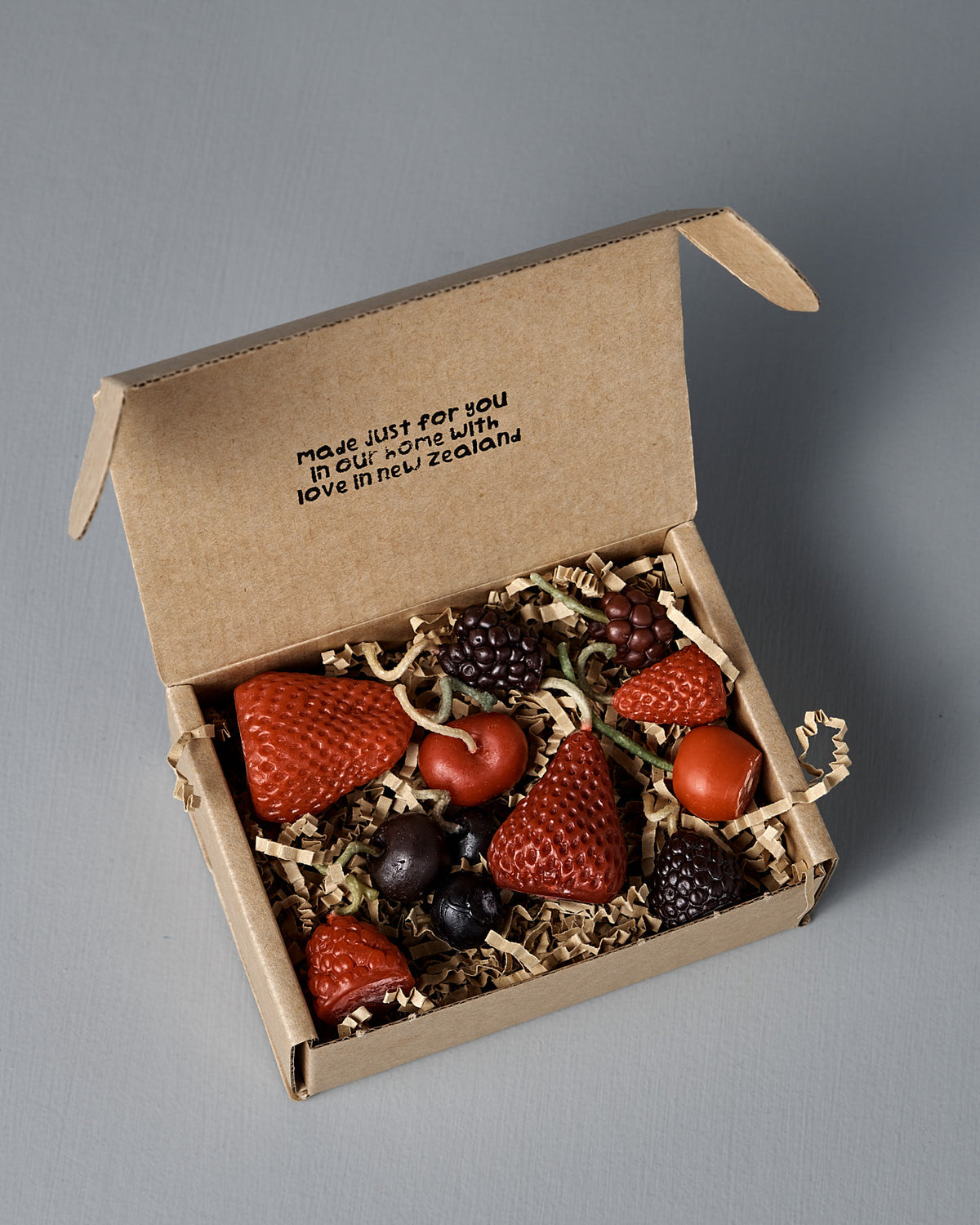 Open cardboard box with an assortment of berry-shaped candles nestled on shredded paper. Text inside lid reads: “made just for you in our home with love in New Zealand.” Perfect as a unique birthday cake decoration, this delightful set of Mixed Berries Candles brings a touch of sweetness to any celebration.