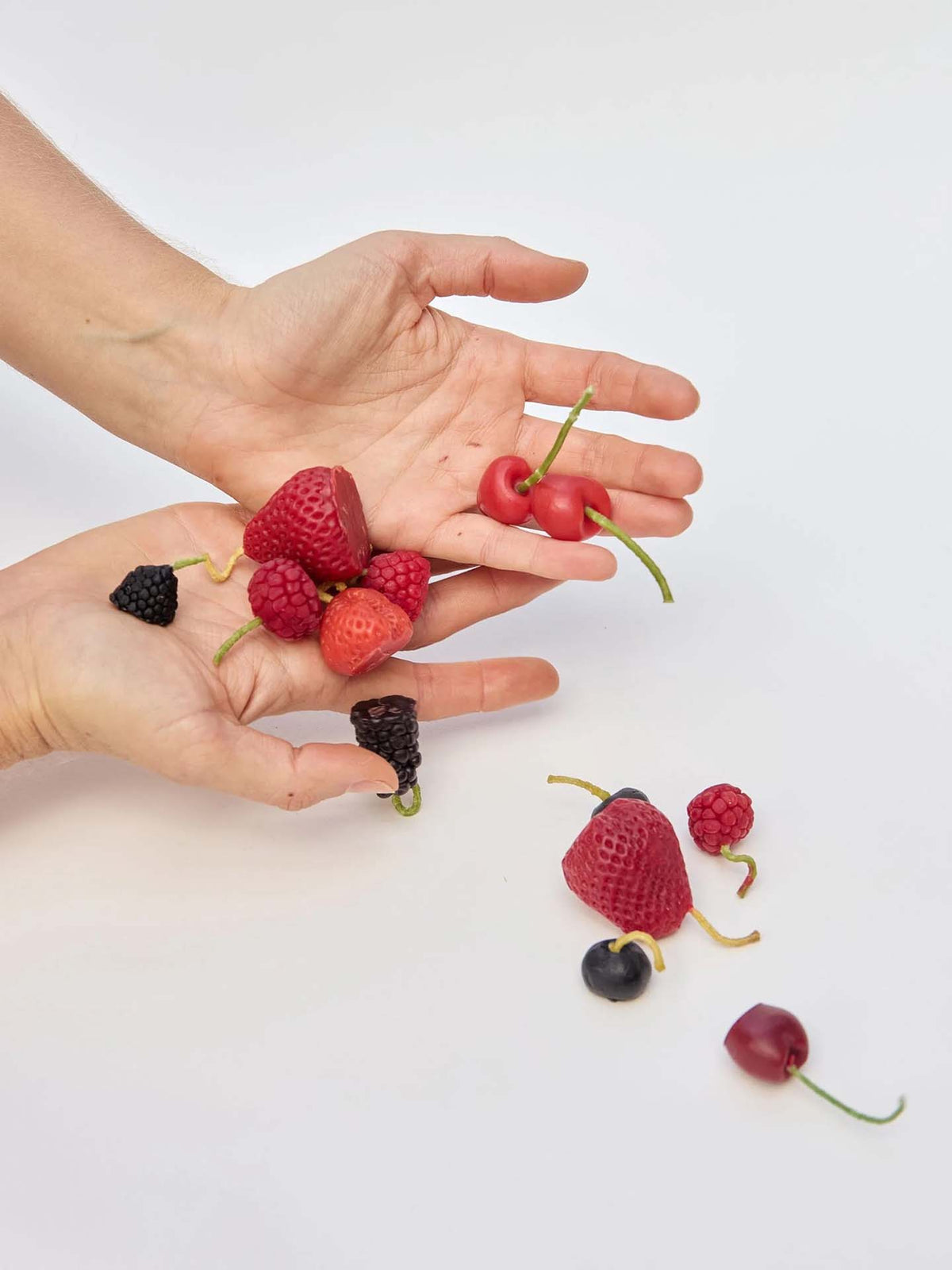Hands holding and releasing strawberries, raspberries, blackberries, and cherries—mirroring the vibrant look of our Mixed Berries Candles – Box of 10—on a white background.