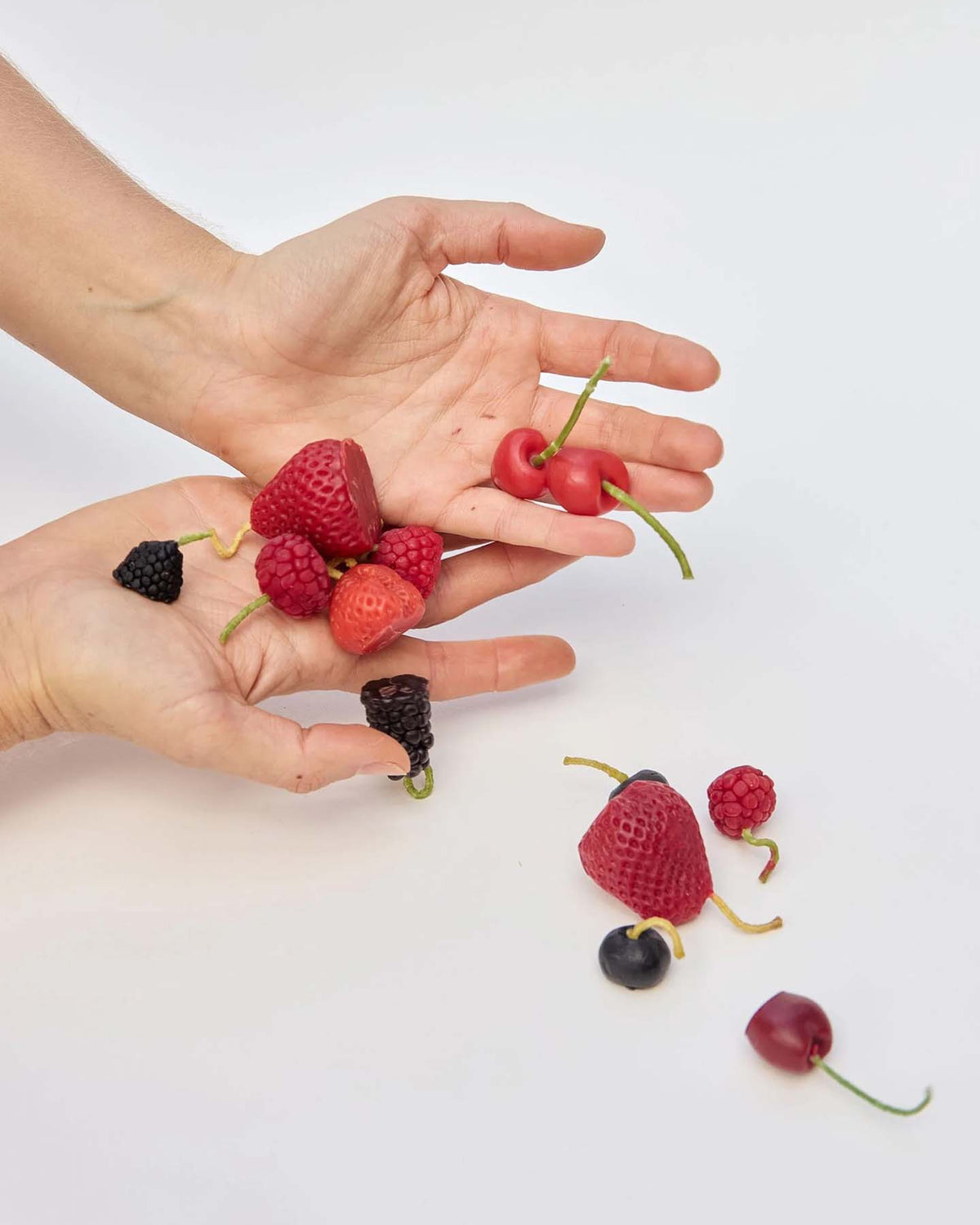 Two hands holding strawberries, cherries, raspberries, blackberries, and blueberries with more mixed berries scattered below on a white background—perfect inspiration for selecting birthday cake decorations or accenting your celebration with Mixed Berries Candles – Box of 12, charming fruit-shaped candles made from New Zealand beeswax.